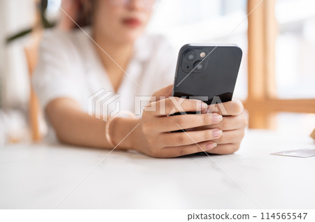 A tired, bored Asian woman leaning on the table and holding her smartphone, waiting for the messages 114565547