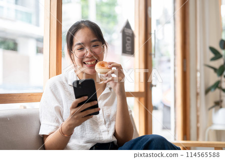 A young, happy Asian woman enjoying a donut while using her smartphone on a sofa in a cafe. 114565588