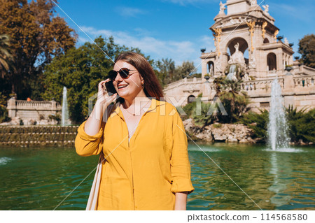 Beautiful stylish woman in trendy wear spending time on street using smartphone. Happy girl talking on the mobile phone near Ciutadella Park in Barcelona. Traveling Europe 114568580