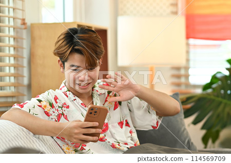 Cheerful young gay male sitting in living room and having video call on mobile phone with their friends Cheerful young gay male sitting in living room and having video call on mobile phone with their friends 114569709