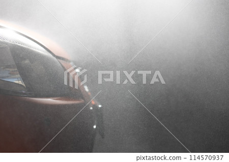 Close-up. Side view. On the headlights and face of the car in shallow patches of water illuminated by the rays of the sun. A beautiful car stands at a car wash on a dark background in the fog. Close-up. Side view. On the headlights and face of the car in shallow patches of water illuminated by the rays of the sun. A beautiful car stands at a car wash on a dark background in the fog. 114570937