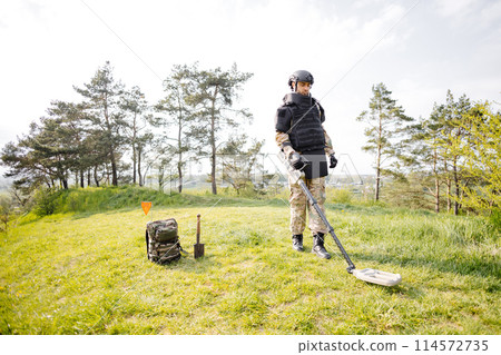 A man in a military uniform and bulletproof vest works in the forest with a metal detector. A minesweeper performs work on demining the territory 114572735