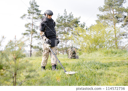 A man in a military uniform and bulletproof vest works in the forest with a metal detector. A minesweeper performs work on demining the territory A man in a military uniform and bulletproof vest works in the forest with a metal detector. A minesweeper performs work on demining the territory 114572736