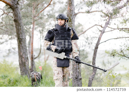 A man in a military uniform and bulletproof vest works in the forest with a metal detector. A minesweeper performs work on demining the territory 114572739