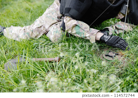 A man in a military uniform and a bulletproof vest works in the forest to demine the territory. A man warns of danger by making a red mark 114572744