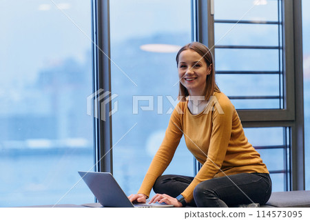 A businesswoman utilizes her laptop while seated by the window of a large corporate building, offering a picturesque view of the city skyline as her backdrop. 114573095