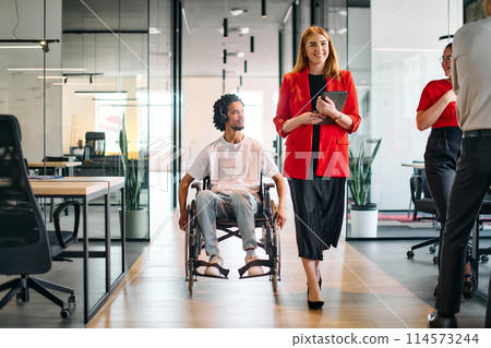 A business leader with her colleague, an African-American businessman who is a disabled person, pass by their colleagues who work in modern offices 114573244