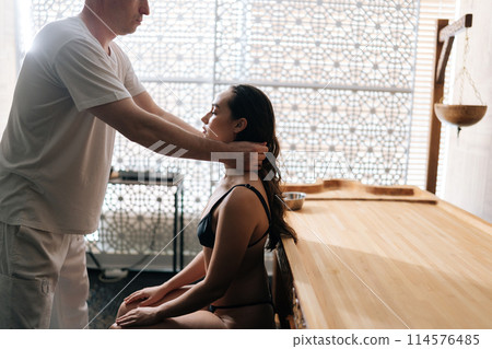 Side view of relaxed female client getting head and neck massage by unrecognizable professional masseur in spa salon with calm interior. Massage therapist male doing massage head and hair for woman. Side view of relaxed female client getting head and neck massage by unrecognizable professional masseur in spa salon with calm interior. Massage therapist male doing massage head and hair for woman. 114576485