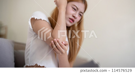 Young woman feeling pain in her elbow while sitting indoors Young woman feeling pain in her elbow while sitting indoors 114576529