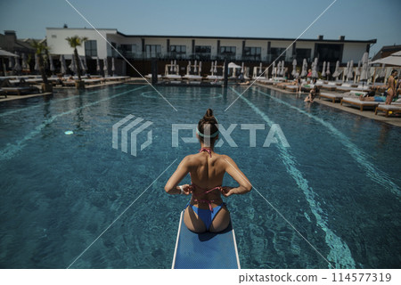 A vibrant lady, dressed in a blue bikini, stands before the pool's cerulean waters. 114577319