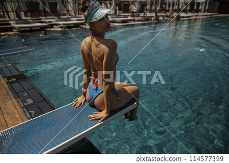 A beautiful young woman in a swimsuit sits by the pool, facing away from the water. A beautiful young woman in a swimsuit sits by the pool, facing away from the water. 114577399