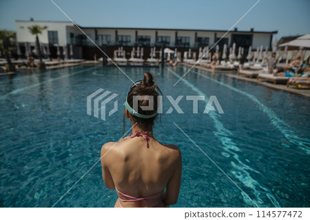 A stunning lady in a bikini reclines by the poolside, her back turned to the water. 114577472