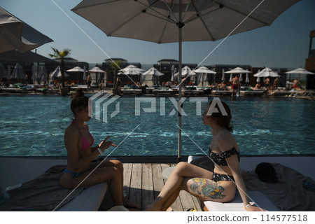 Two girlfriends in bikinis lounge poolside, enjoying the warmth of the sun. Two girlfriends in bikinis lounge poolside, enjoying the warmth of the sun. 114577518