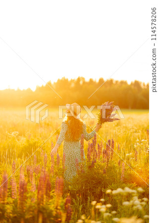 A young woman in a stylish green dress and hat enjoys the sunset in a field with lupins. A young woman in a stylish green dress and hat enjoys the sunset in a field with lupins. 114578206