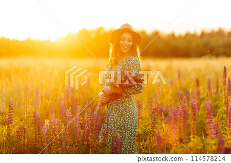 A young woman in a stylish green dress and hat enjoys the sunset in a field with lupins. 114578214