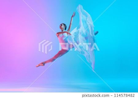 Ballet dancer performs jumping split in motion with flowing white cloth in neon light against vivid gradient background. Ballet dancer performs jumping split in motion with flowing white cloth in neon light against vivid gradient background. 114578482