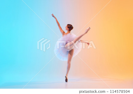Rear view of young ballerina in white tutu doing arabesque in motion in neon light against blue-orange gradient background. 114578504