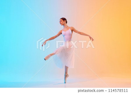Young ballerina in white tutu an pointe practicing dance moves in neon light against blue-orange gradient background. Young ballerina in white tutu an pointe practicing dance moves in neon light against blue-orange gradient background. 114578511