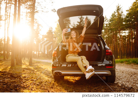 Stylish woman takes selfie, communicates via video call from smartphone in trunk of a car at sunset. Stylish woman takes selfie, communicates via video call from smartphone in trunk of a car at sunset. 114579672