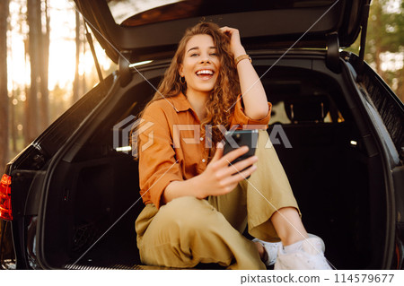 Young female tourist in casual clothes sits in the trunk of a car and uses a phone. Adventure. Young female tourist in casual clothes sits in the trunk of a car and uses a phone. Adventure. 114579677