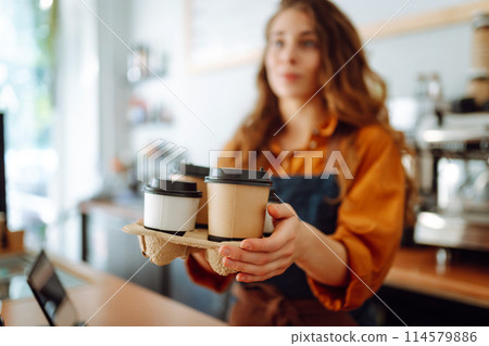 Cheerful woman in an apron at the bar counter holds coffee glasses in a cafe. Takeaway food concept. Cheerful woman in an apron at the bar counter holds coffee glasses in a cafe. Takeaway food concept. 114579886