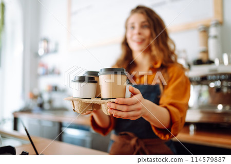 Cheerful woman in an apron at the bar counter holds coffee glasses in a cafe. Takeaway food concept. Cheerful woman in an apron at the bar counter holds coffee glasses in a cafe. Takeaway food concept. 114579887