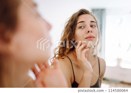 Portrait of a young woman touching pimple on her face while looking in mirror. Facial skin problems. 114580064