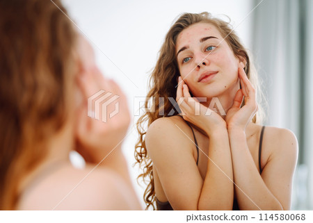 Portrait of a young woman touching pimple on her face while looking in mirror. Facial skin problems. 114580068