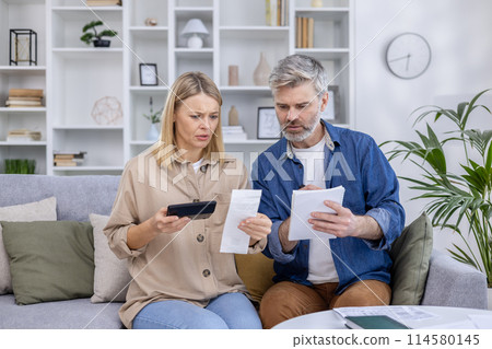 Worried mature couple reviewing bills together at home, calculating finances with a smartphone and notepad. Concept of financial planning and family budgeting. Worried mature couple reviewing bills together at home, calculating finances with a smartphone and notepad. Concept of financial planning and family budgeting. 114580145