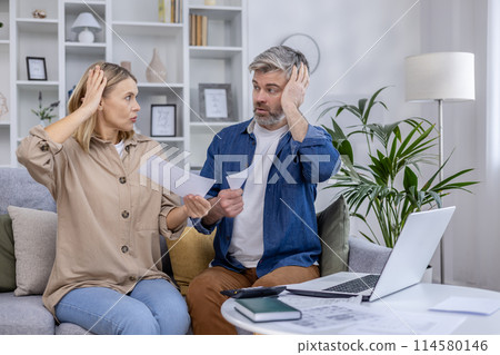 Stressed couple sitting on the couch discussing bills and finances. They appear worried while holding papers in their hands and looking at each other. Stressed couple sitting on the couch discussing bills and finances. They appear worried while holding papers in their hands and looking at each other. 114580146