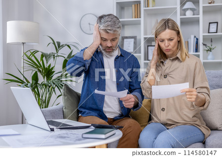 Worried couple sitting on the couch while reviewing financial documents, experiencing financial stress and difficulties Worried couple sitting on the couch while reviewing financial documents, experiencing financial stress and difficulties 114580147