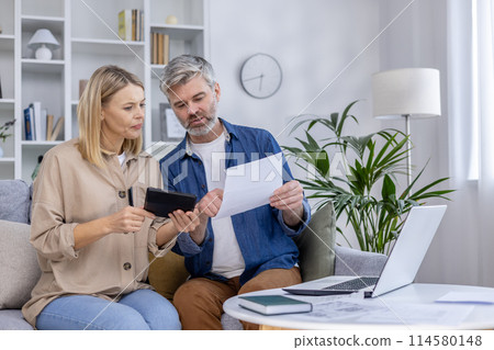 Mature couple sitting on a couch, reviewing documents and using a tablet at home. They seem focused and engaged in a financial discussion. 114580148