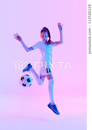 Full-length image of teen girl in white uniform, soccer player in motion, playing against pink studio background in neon light 114580226