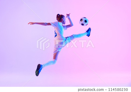 Dynamic image of focused teen girl, soccer player in motion, practicing, kicking ball in jump against pink studio background in neon light 114580230