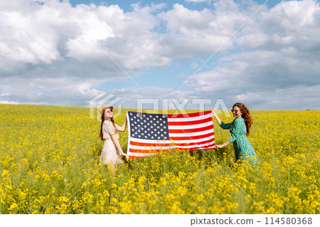 Two Young woman with american flag on blooming meadow. 4th of July. USA flag fluttering in the wind. Two Young woman with american flag on blooming meadow. 4th of July. USA flag fluttering in the wind. 114580368