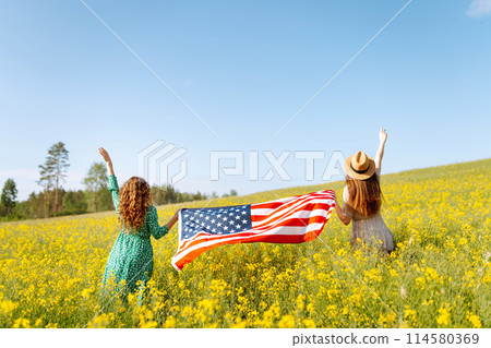 Two Young woman with american flag on blooming meadow. 4th of July. USA flag fluttering in the wind. Two Young woman with american flag on blooming meadow. 4th of July. USA flag fluttering in the wind. 114580369