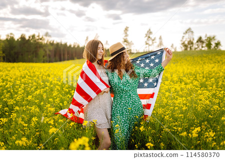 Two Young woman with american flag on blooming meadow. 4th of July. USA flag fluttering in the wind. 114580370