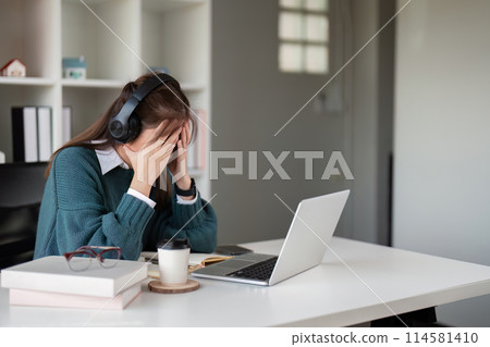 Young woman feeling stressed while wearing headphones and using a laptop in a home office Young woman feeling stressed while wearing headphones and using a laptop in a home office 114581410
