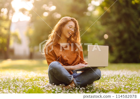 Portrait of young beautiful female freelancer sitting on green meadow with laptop. Education online. 114582078