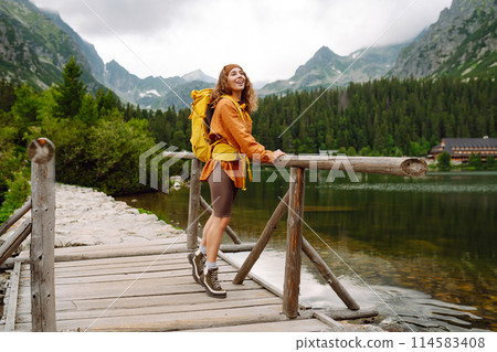 Female traveler with yellow hiking backpack against the backdrop of mountain lake with hiking poles. 114583408