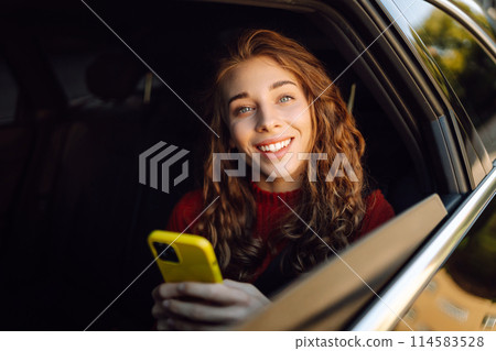 Positive young woman in casual clothes sits in the back seat of a car with a mobile phone. Positive young woman in casual clothes sits in the back seat of a car with a mobile phone. 114583528