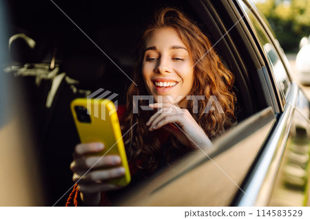 Positive young woman in casual clothes sits in the back seat of a car with a mobile phone. 114583529