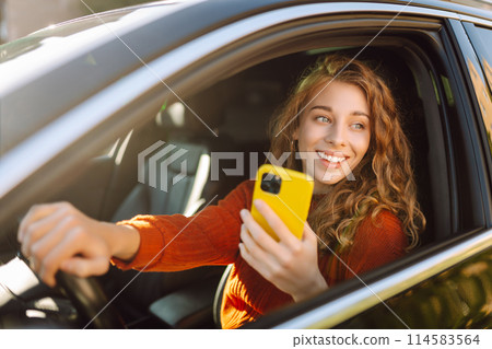 Portrait of a young woman sitting in a car in the driver's seat looking into a smartphone. 114583564