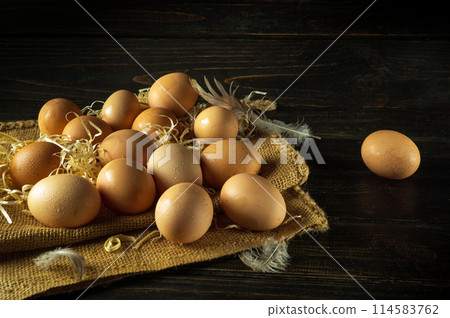 Eggs stacked on burlap with straw before harvesting. Concept of preparing eggs for cooking. Place for advertising on a dark background Eggs stacked on burlap with straw before harvesting. Concept of preparing eggs for cooking. Place for advertising on a dark background 114583762
