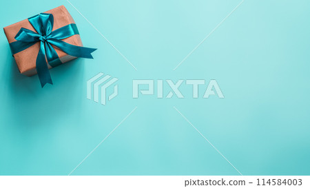 Gift box in craft wrapping paper and green satin ribbon on turquoise blue background, copy space right. Beautiful Christmas, New Year or Birthday present, flat lay or top view. Banner Gift box in craft wrapping paper and green satin ribbon on turquoise blue background, copy space right. Beautiful Christmas, New Year or Birthday present, flat lay or top view. Banner 114584003
