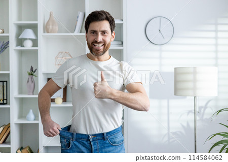 Cheerful man displaying weight loss success by holding out loose jeans and giving a thumbs up, standing in a modern living room. 114584062