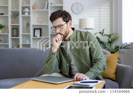 Thoughtful young man working on his laptop at home. He is thinking and planning while sitting on the sofa. Thoughtful young man working on his laptop at home. He is thinking and planning while sitting on the sofa. 114584066
