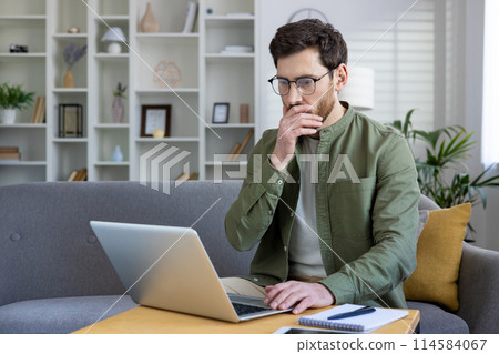 Man working from home on a laptop, thinking and analyzing projects in a modern living room setting. 114584067