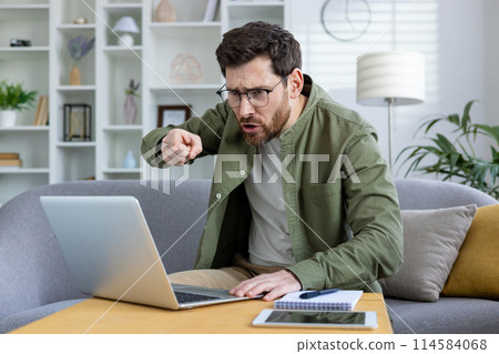 Angry man in glasses and green shirt pointing at laptop during a video call, expressing frustration in a home office setting. Angry man in glasses and green shirt pointing at laptop during a video call, expressing frustration in a home office setting. 114584068
