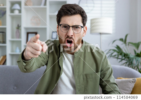 Angry man wearing glasses and a green shirt pointing and yelling while sitting on a couch in a modern living room with shelves ,in the background. 114584069